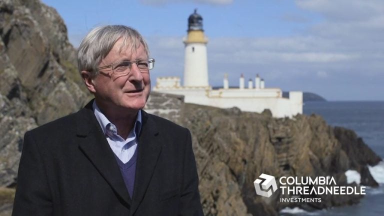 A gray haired man with glasses against the background of a lighthouse